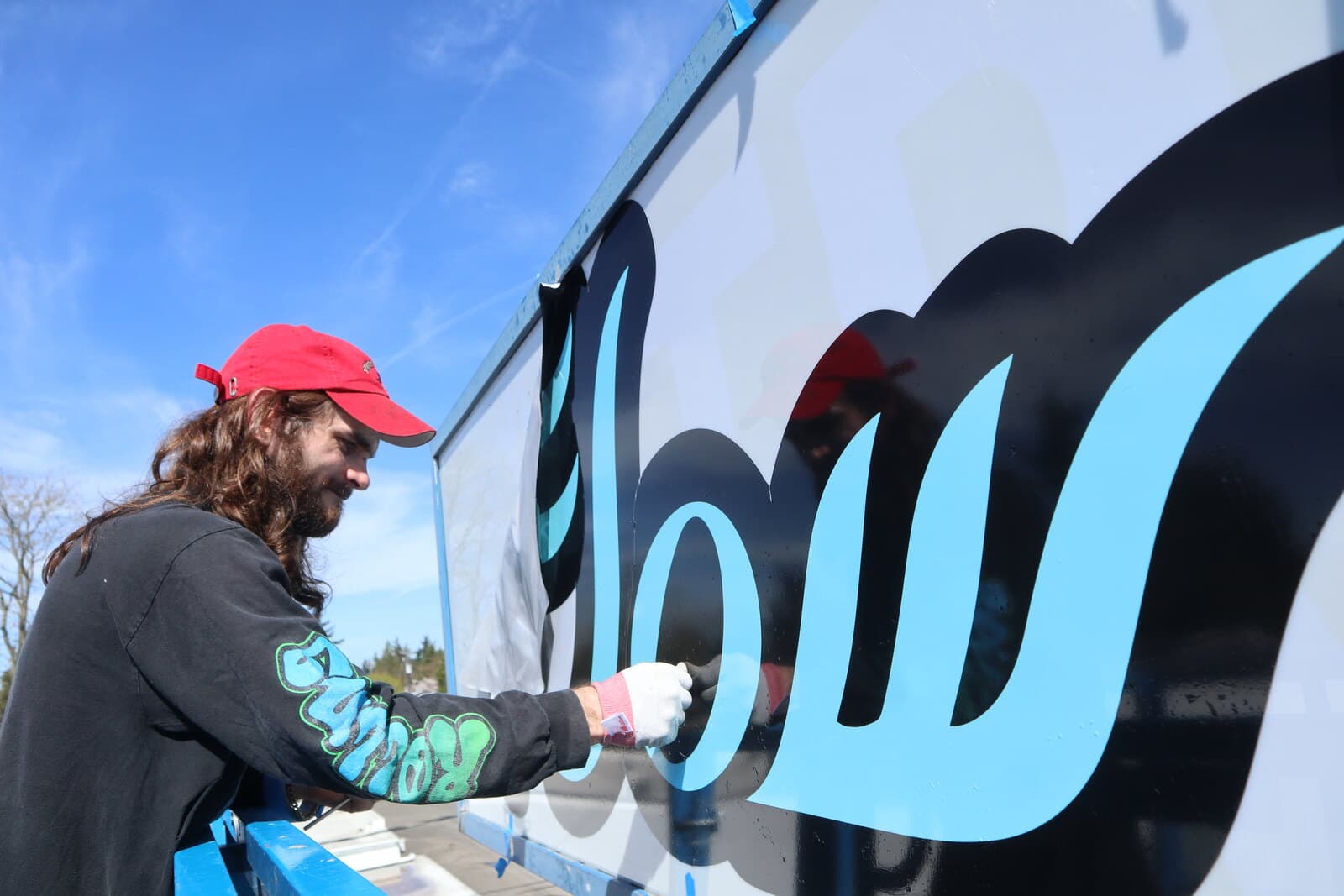 A man on a lift smoothing cut vinyl lettering onto a large sign face by hand, under a blue sky